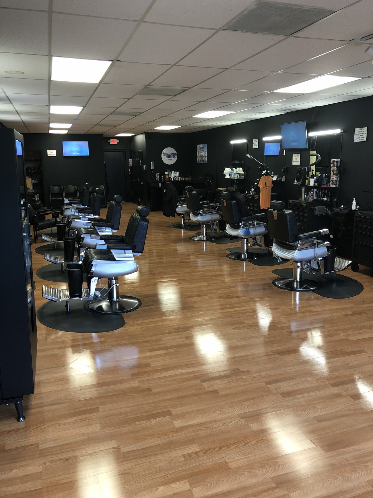 Inside the shop looking toward the back: two rows of black leather barber chairs on chrome bases facing mirrored stations along black walls, with a polished light-wood floor and TVs mounted overhead.